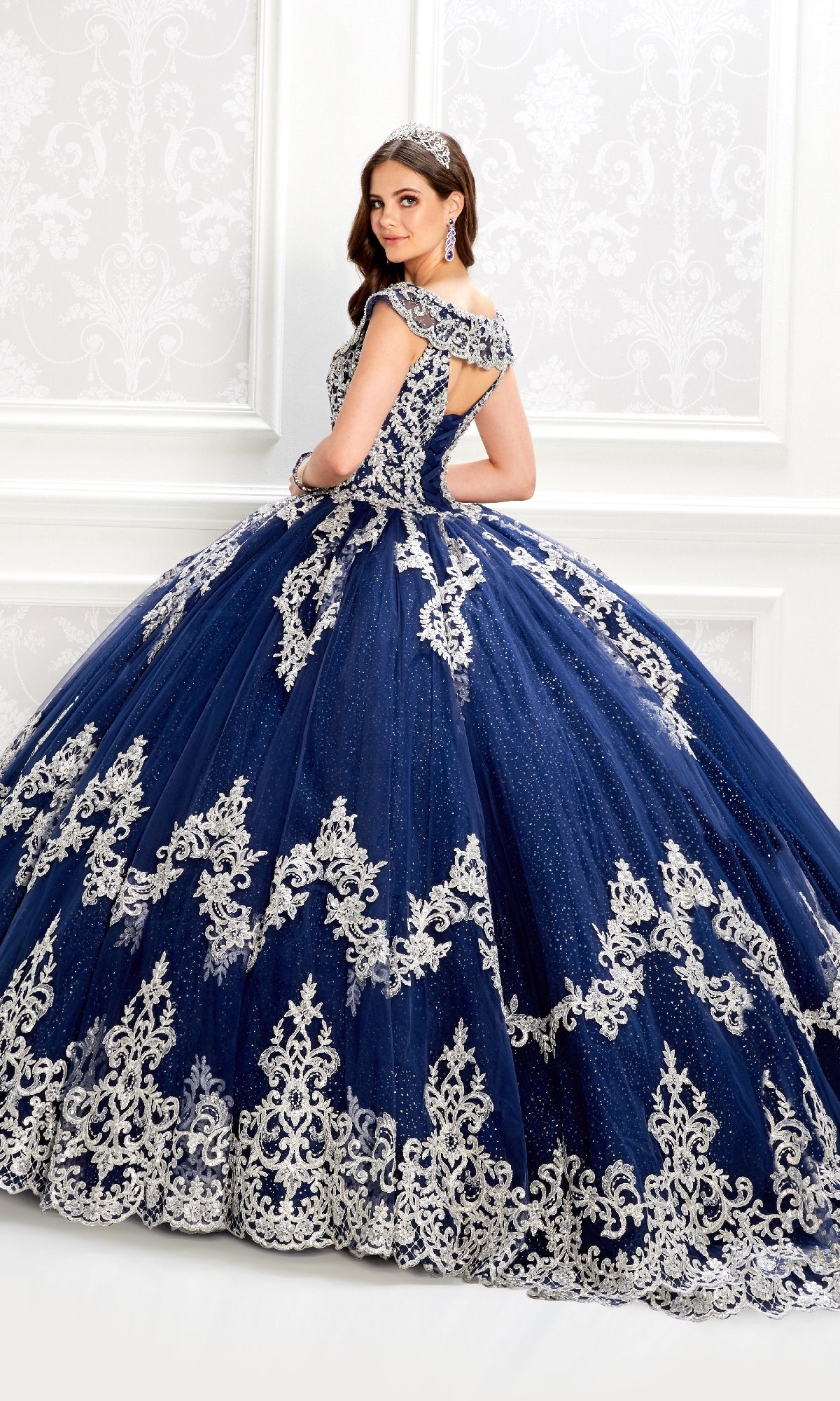 Woman wears the Quinceanera Dress PR22035 By Princesa, a dark blue ball gown with a glitter tulle skirt and silver embroidery, posing in an elegant white room while looking over her shoulder and wearing a jeweled headband.